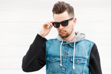 Yakışıklı hipster adam bir kot ceket ve beyaz bir ahşap duvar yakınındaki siyah güneş gözlüğü