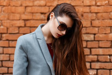 Güzel genç kız güneş gözlüğü ve gri ceket tuğla duvara yakın aşağı görünüyor