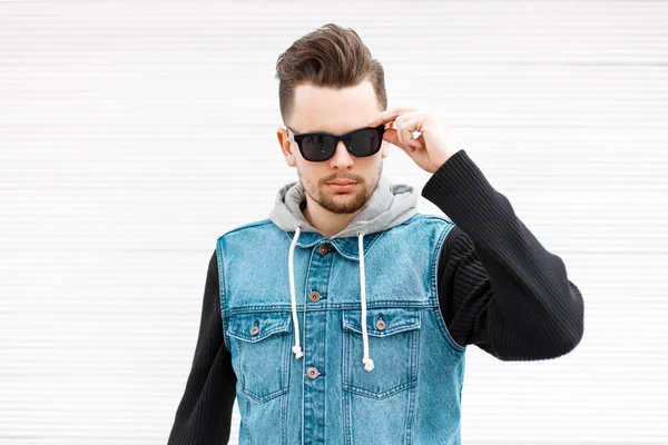 Handsome stylish man with sunglasses in a jeans jacket near a vintage white wooden wall — Stock Photo, Image