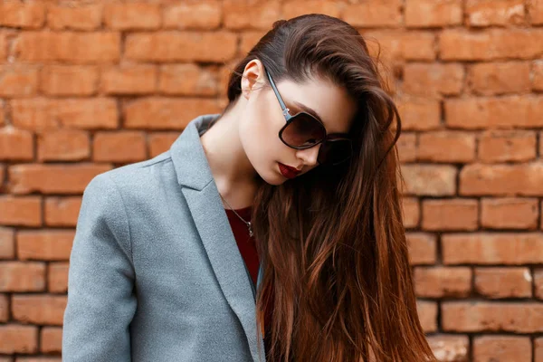 Schönes junges Mädchen mit Sonnenbrille und grauem Mantel blickt nahe Ziegelmauer hinunter — Stockfoto