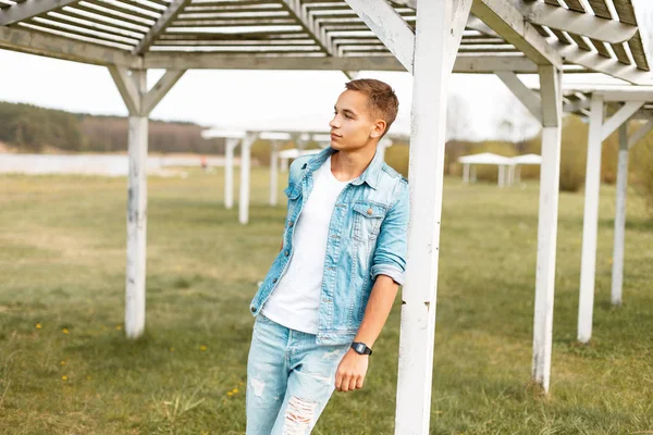 Jovem bonito com jeans rasgados e uma jaqueta de ganga perto de um dossel de praia de madeira branca ao ar livre — Fotografia de Stock