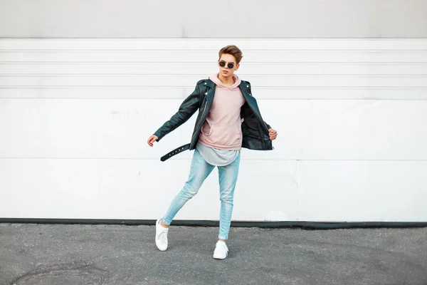 Knappe jonge trendy man met zonnebril in een zwart lederen jas, een roze trui, blauw merk jeans en witte schoenen in beweging — Stockfoto