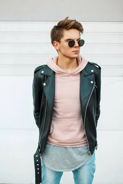 Guapo joven hombre de moda con un corte de pelo en gafas de sol en una chaqueta de cuero negro y una sudadera rosa cerca de una pared de metal blanco —  Fotos de Stock