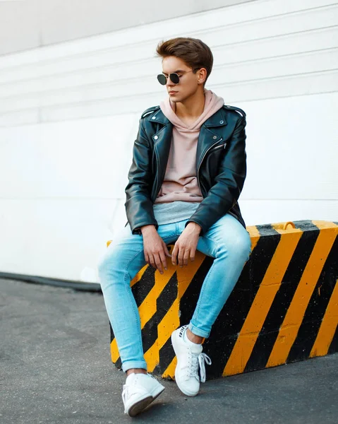 Молодий красивий чоловік з сонцезахисними окулярами в стильній чорній шкіряній куртці, рожевий светр, сині джинси і біле взуття сидить на чорно-жовтій плиті — стокове фото