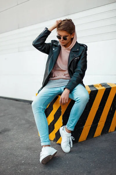 Jovem elegante bonito com óculos de sol em uma jaqueta de couro preto, jeans azuis e tênis brancos coloca um capuz sentado em um preto-e-amarelo — Fotografia de Stock