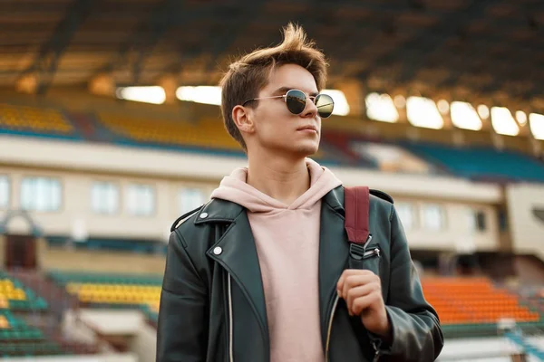 Beau portrait d'un jeune sportif avec des lunettes de soleil dans une veste noire avec un sac à dos au stade — Photo