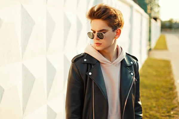 Joven guapo con gafas de sol con estilo en una chaqueta de cuero, una sudadera rosa cerca de una pared blanca en un día soleado — Foto de Stock