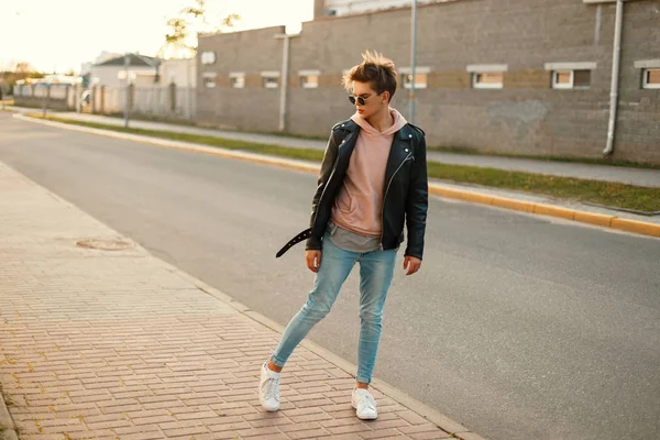 Jovem bonito com óculos de sol em uma jaqueta de couro preto, jeans azuis e sapatos brancos na rua perto da estrada — Fotografia de Stock