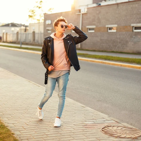 Bonito jovem elegante com óculos de sol vintage em uma jaqueta de couro e sapatos brancos ao pôr do sol — Fotografia de Stock