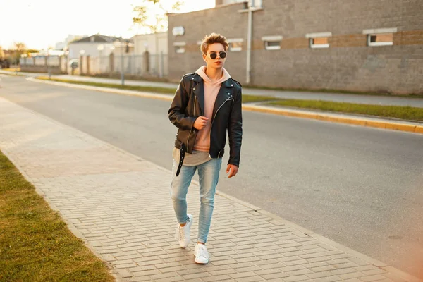 Pohledný mladý muž s účes v sluneční brýle v kožené bundy, džíny a bílé tenisky na ulici při západu slunce — Stock fotografie