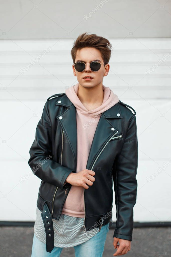 Young handsome man in a black branded leather jacket and a pink sweatshirt near a white metal wall