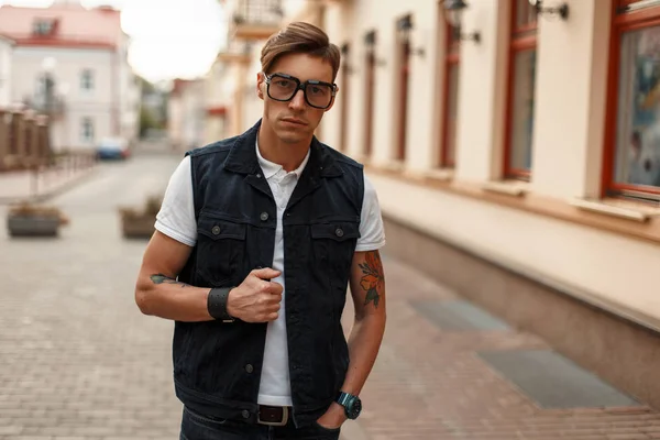 Joven hombre guapo en gafas con estilo en chaleco vaquero vintage y una camisa blanca cerca del edificio — Foto de Stock