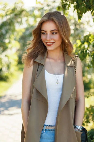 Zadowolony, młoda kobieta z białym uśmiechem w modne ciuchy z torebka spaceru w parku w słoneczny dzień wiosny — Zdjęcie stockowe