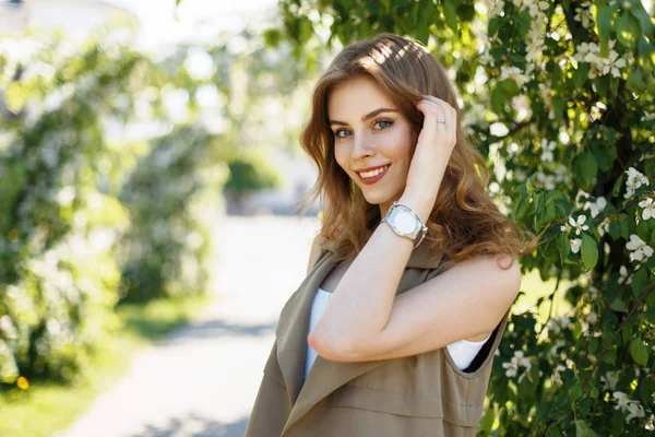 Hermosa mujer feliz con una sonrisa en ropa de moda con un reloj cerca de árboles florecientes en un día soleado de primavera —  Fotos de Stock