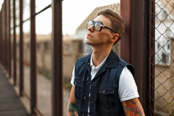 Yakışıklı genç adam gözlüklü bir metal ızgara yakınında modern giyimleri
