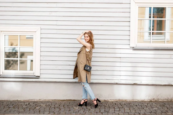 Красивая молодая женщина улыбается в стильной одежде, джинсах и черных туфлях с сумочкой рядом с деревянным деревянным домом с окнами — стоковое фото