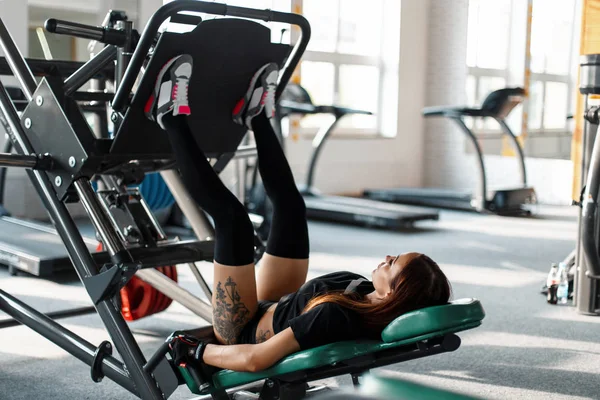 Sexig kvinna med en tatuering i sport underkläder pumpar fötterna på en sport maskin i gymmet — Stockfoto