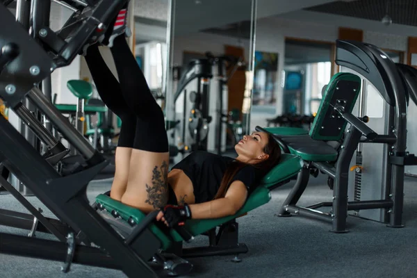 Νεαρή γυναίκα με ένα τατουάζ εκπαιδεύει τα πόδια της σε ένα προσομοιωτή σπορ στο γυμναστήριο — Φωτογραφία Αρχείου