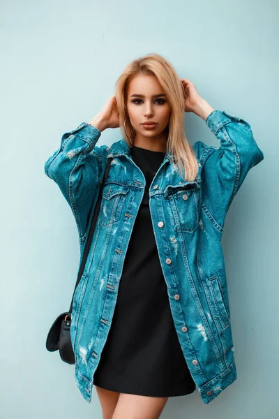 Schöne blonde Frau mit Handtasche in blauer Jeanskleidung posiert in der Nähe der blauen Wand — Stockfoto