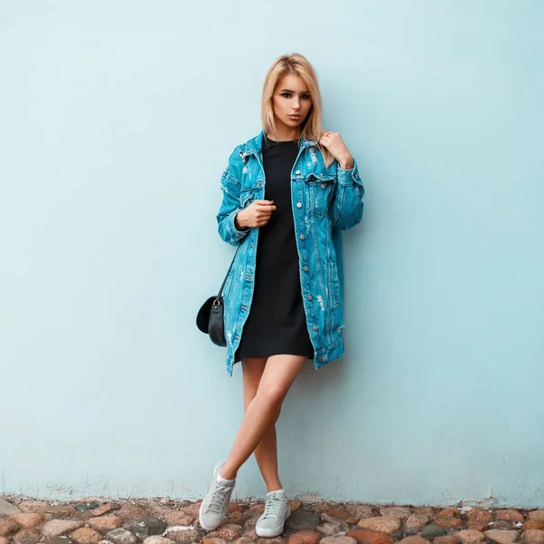 Elegante hermosa mujer con un bolso de mano en ropa de mezclilla de moda con zapatillas de deporte cerca de una pared azul —  Fotos de Stock