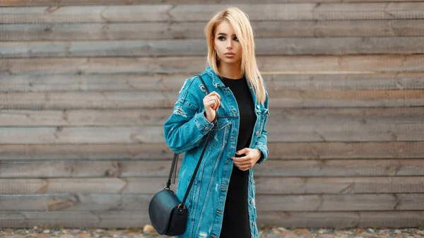 Schöne junge blonde Frau mit einer Handtasche in Jeansjacke in der Nähe einer hölzernen Vintage-Wand — Stockfoto