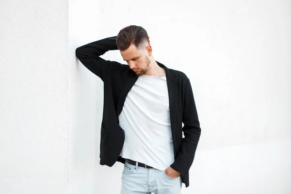 Homem bonito na moda em casaco elegante preto posando perto da parede branca — Fotografia de Stock