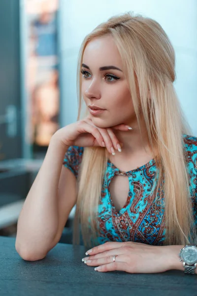 Portret van een mooie jonge blonde vrouw in een blauwe jurk met een patroon zit in een café op de straat — Stockfoto