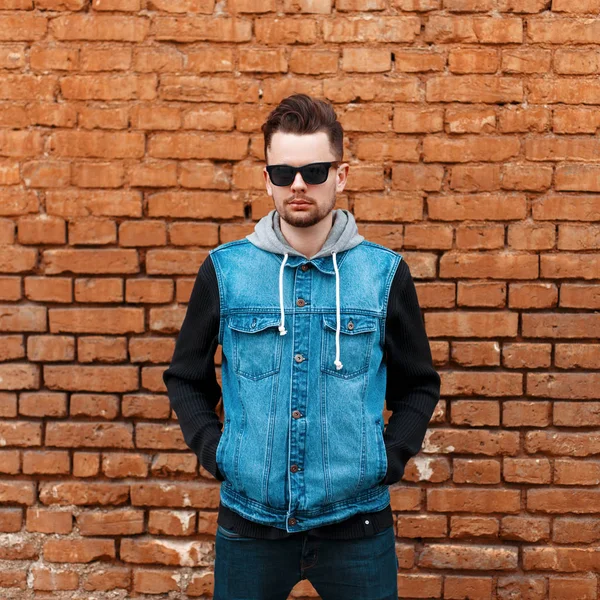 Handsome hipster guy in a denim jacket near a brick wall — Stock Photo, Image