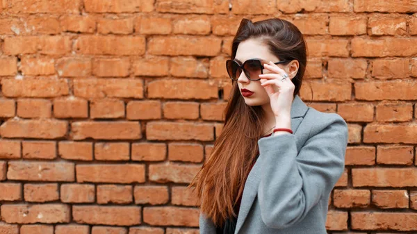 Стильная молодая красивая женщина в солнечных очках в сером пальто возле кирпичной стены — стоковое фото