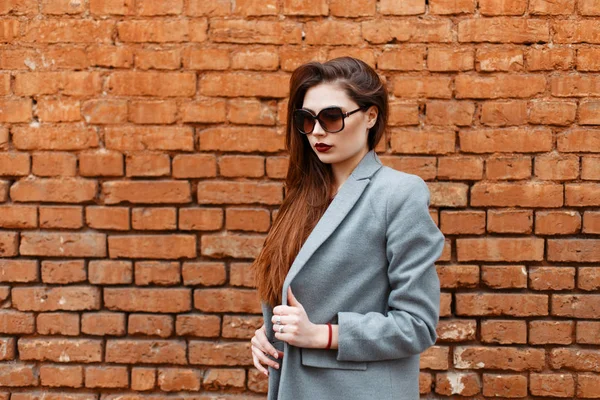 Belle fille brune modèle dans un manteau gris avec des lunettes de soleil près d'un mur de briques rouges — Photo