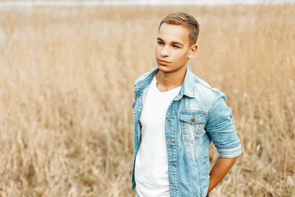 Guapo joven con estilo en una chaqueta jeans y una camisa blanca en la naturaleza en una hierba gris — Foto de Stock