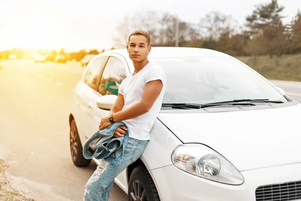 Handsome young man in a white tank top near the car at sunset. Traveling on the roads — Stock Photo, Image
