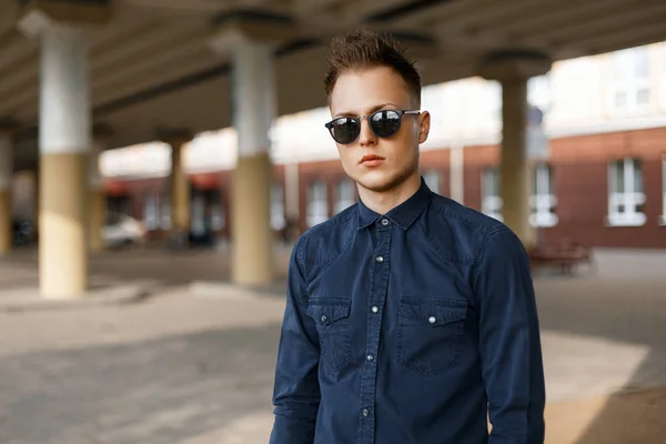 Joven hombre guapo con estilo en gafas de sol y una camisa azul en un día soleado en la calle — Foto de Stock