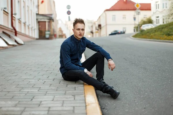 Handsome young guy in a blue shirt and black sneakers sits on the asphalt on the street — Stock Photo, Image