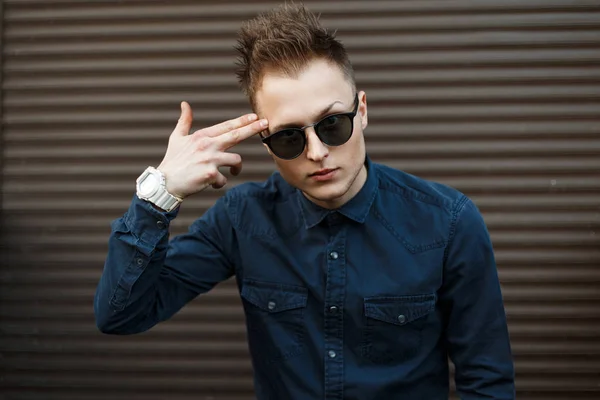 Schöner junger Mann mit Sonnenbrille und blauem Hemd zeigt eine Pistole aus seinen Fingern in der Nähe der Metallwand — Stockfoto