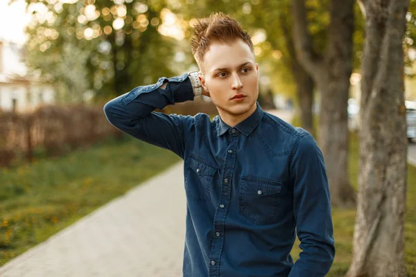 Красивый молодой стильный мужчина в синей рубашке позирует в парке в солнечный день — стоковое фото