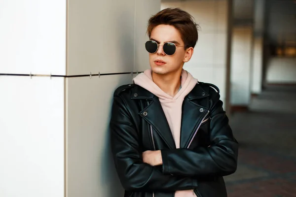 Przystojny, młody facet z okulary w modne markowe kurtki stoi w pobliżu białej ścianie — Zdjęcie stockowe