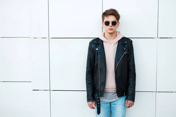 Hombre joven con una chaqueta de cuero negro y una sudadera rosa cerca de una pared blanca. Aviador de gafas de sol vintage — Foto de Stock