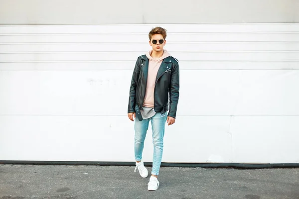 Stijlvolle knappe jonge man met zonnebril in een zwart lederen jas, blue Trendy jeans en witte trendy sneakers in de buurt van een muur wit metaal — Stockfoto