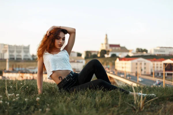 Mujer joven en una camiseta blanca está descansando y sentado en la hierba disfruta del momento, fondo urbano — Foto de Stock