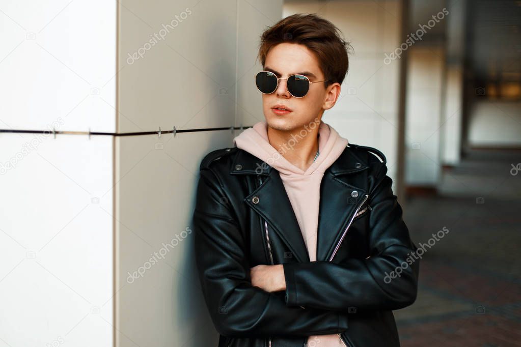 Handsome young guy with sunglasses in a fashionable branded jacket stands near a white wall