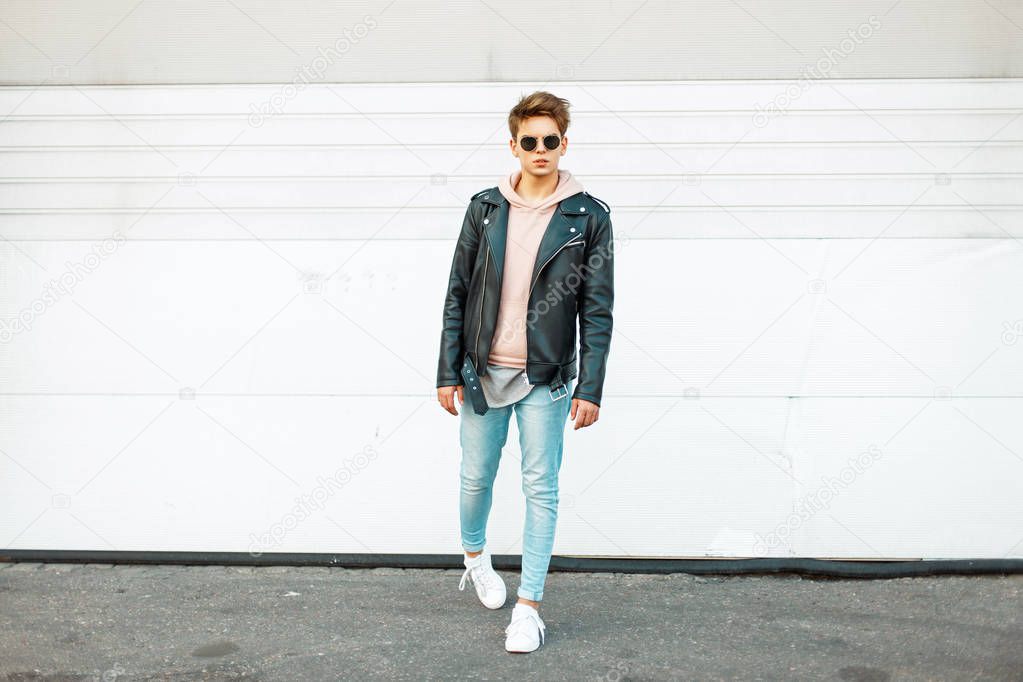 Stylish handsome young man with sunglasses in a black leather jacket, blue fashionable jeans and white trendy sneakers near a white metal wall