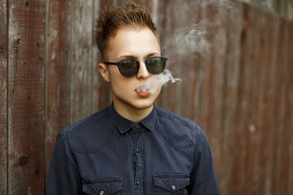 Young stylish man in sunglasses with smoke smokes a cigarette near a wooden wall — Stock Photo, Image