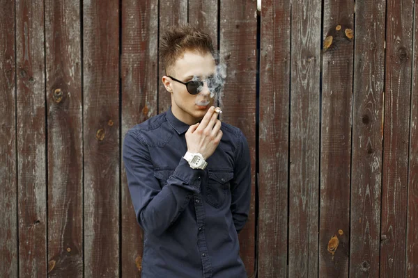 Joven con gafas de sol lanza humo y fuma un cigarrillo cerca de una pared de madera — Foto de Stock