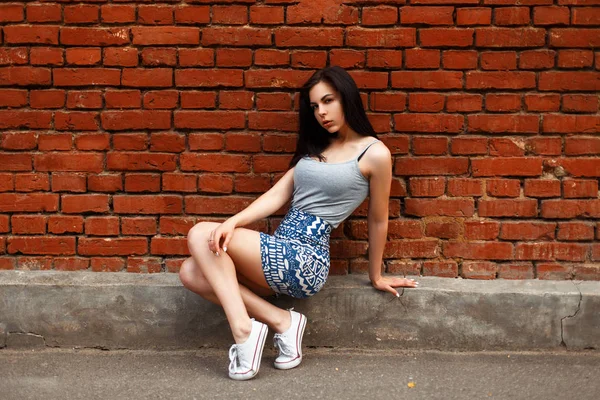 Mooi jong meisje in een zomer rok en grijs T-shirt met sneakers zit in de buurt van een rode bakstenen muur — Stockfoto