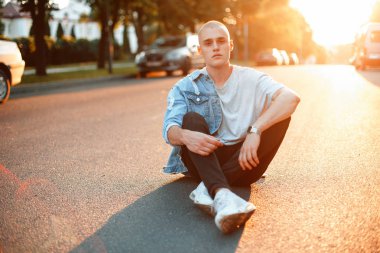 Yakışıklı genç gençlik adam şık şık denim giysiler içinde asfalt yolda günbatımında oturur.