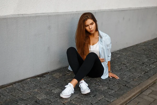 Mooie stijlvolle jong meisje in trendy casual casual kleding, zittend op een tegel in de buurt van een muur — Stockfoto