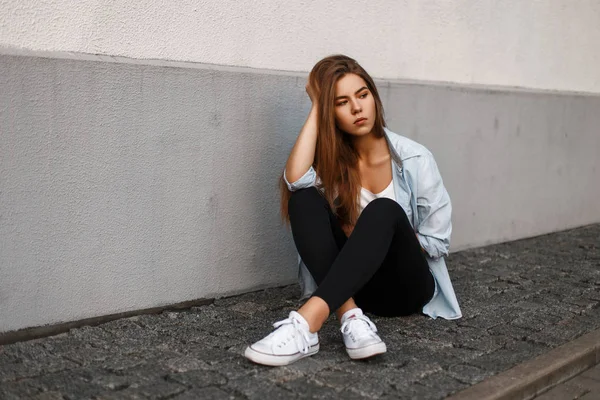 Güzel genç Amerikalı manken kadın siyah pantolon ve beyaz ayakkabı duvara yakın oturan mavi gömlekli