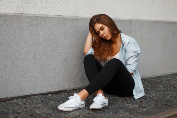 Hermosa mujer de moda en camisa azul casual y zapatos blancos sentados en baldosas cerca de la pared — Foto de Stock
