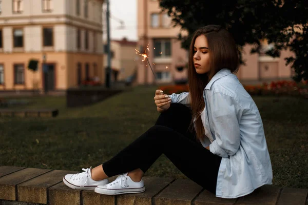 Belle fille élégante à la mode dans un style décontracté dans une chemise, gtans noirs et chaussures blanches avec le feu du Bengale se trouve près d'une pelouse dans la rue — Photo